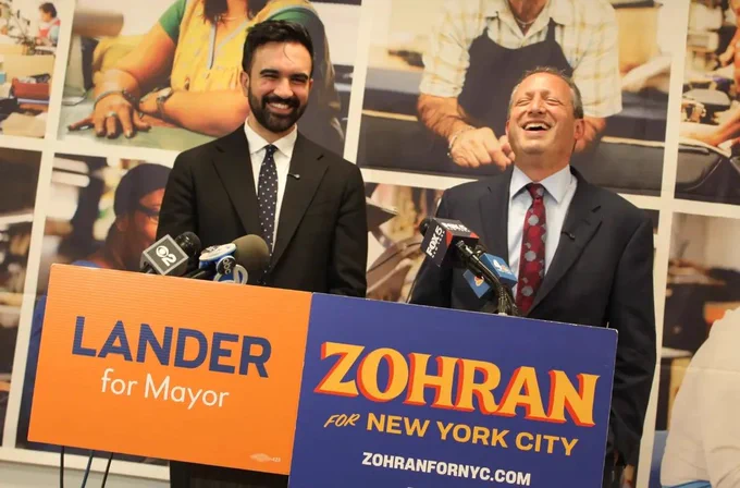 A photograph of Zohran Mamdani and Brad Lander at their co-endorsement press conference. Mamdani appears behind a Lander for Mayor sign and Lander appears behind a Zohran for New York City sign.