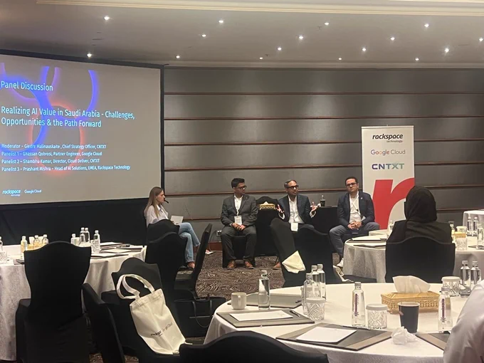 Four panelists are seated in front of a projected presentation titled "Realizing AI Value in Saudi Arabia - Challenges, Opportunities & the Road Ahead" at an event sponsored by entities such as Rackspace, Google Cloud, and CNTXT. The audience in the foreground listens attentively.
