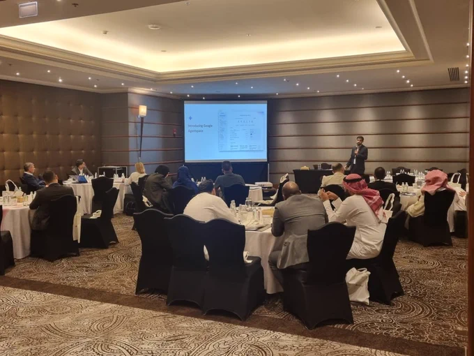 A person is giving a presentation in a conference room with an audience seated at tables. The screen displays graphs and charts. The setting appears professional and well-attended.