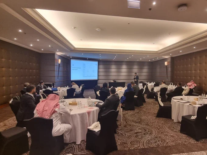 Individual presenting a PowerPoint slide in a conference room filled with attendees seated at round tables.