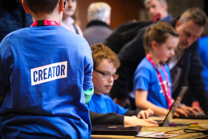 The back our a young person wearing a blue t shirt that says 'creator' on the back. There are other creators sat at laptops blurred in the background.
