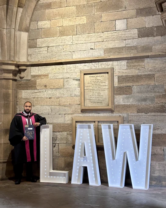A law graduate in black and magenta academic robes stands the university cloisters beside large illuminated letters spelling "LAW". He holds a graduation certificate folder that reads "Graduation" in gold lettering.