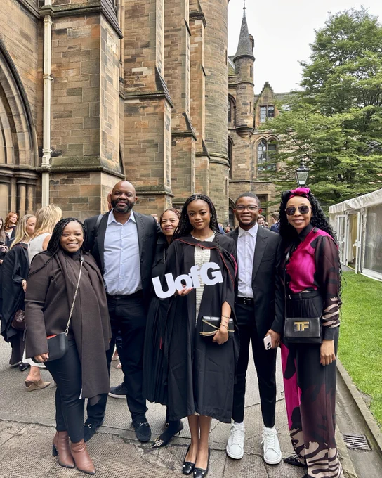 A family of six people, including a graduating student in black robes holding a "UofG" sign, stand together smiling outside the University cloisters.
