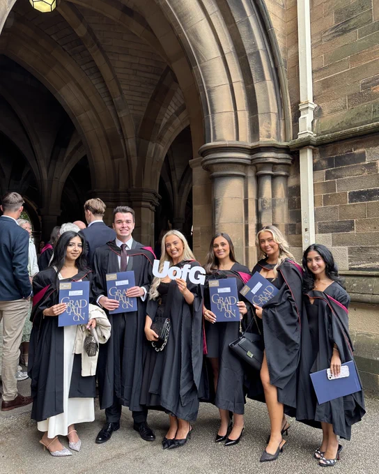 A group of six graduates stand together by the cloisters with their graduation booklets and a 'UofG' sign.