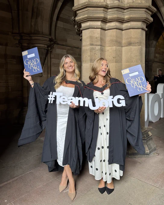  Two graduates in black gowns stand smiling in the cloisters, holding graduation folders and a "#TeamUofG" sign. 
