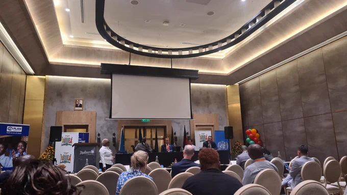 A conference room with speakers seated at the front and delegates seated watching. 