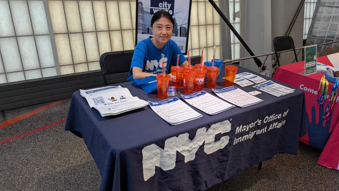 A staff member is sitting at a table with a table cloth, papers, and cups for visitors to take. She is sitting next to another table.