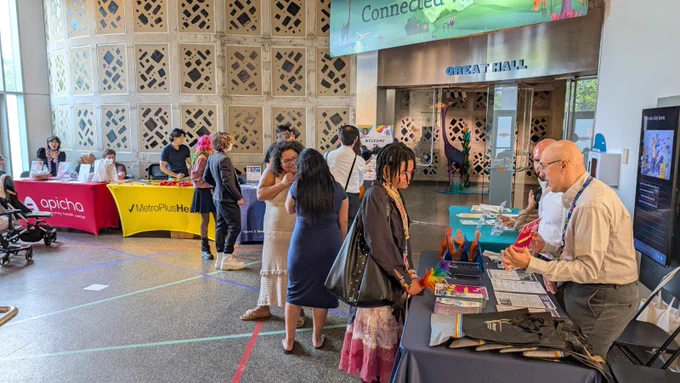 A resource fair is taking place inside a lobby of a museum with many people visiting tables to collect resources and promotional materials. A man at one table is holding up a flyer while explaining it to a visitor.
