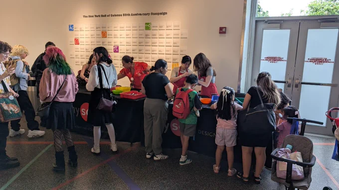 A resource fair is taking place inside a lobby of a museum with many people visiting tables to collect resources and promotional materials. Children stand with their parents who are looking at the resources.