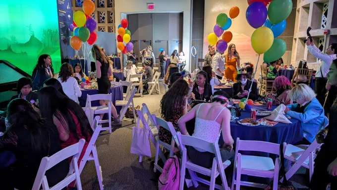 Youth and families are sitting down at tables decorated with colorful balloons for a ceremony inside a building. A photographer is taking pictures of attendees. 