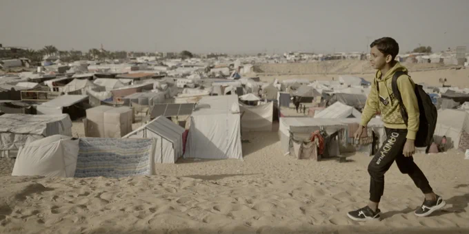 a boy with wearing a a backpack is walking along a sandy path - in the background we can see a village of tents and temporary accommodation build on the sandy ground in Gaza
