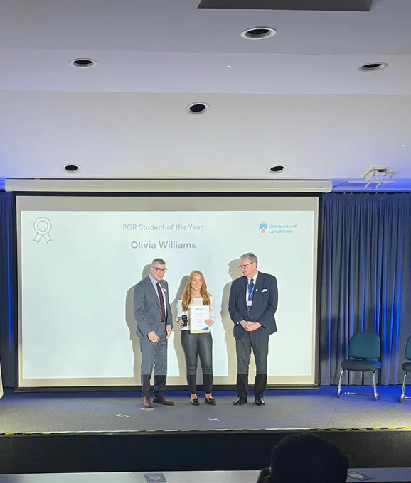Olivia Williams stood on stage holding a certificate and plaque with Professor Graham Baldwin and Professor StJohn Crean. Behind them is a screen which states PGR Student of the Year Olivia Williams.