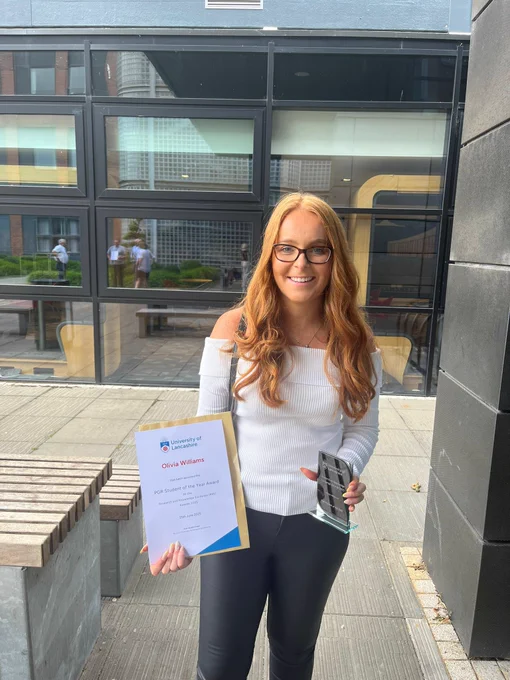 Olivia Williams stood holding a certificate titled PGR student of the year and a mirrored plaque.