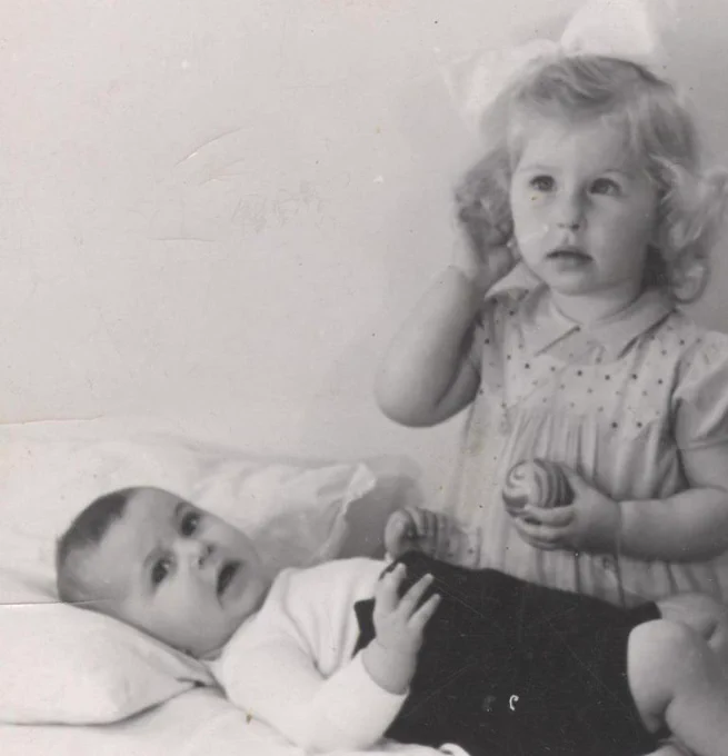 A black and white photo of a toddler sitting next to a baby who is lying down, both looking curiously towards something out of frame.