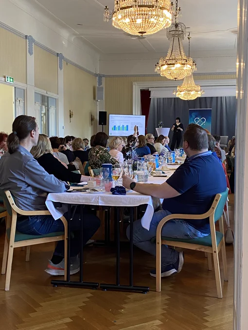 Kuva pöytien ääressä istuvasta yleisöstä kuuntelemassa asiantuntijapuheenvuoroa