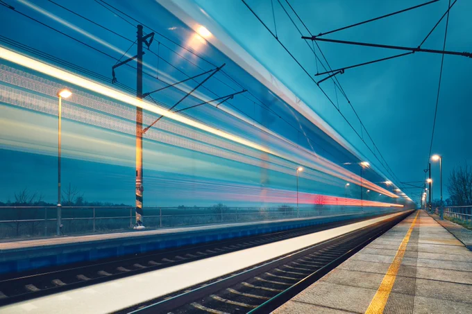 A long exposure photograph capturing the blurred motion of a train passing through a station at twilight, with vibrant streaks of light reflecting the train's speed and illuminated overhead power lines against a dusky sky.