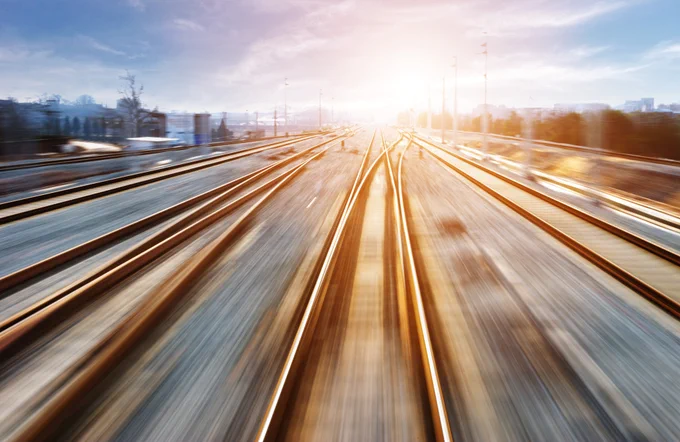 Stock image of sunlit railway tracks converging in the distance, with a blurred motion effect to suggest high speed, set against a bright sky and urban horizon.