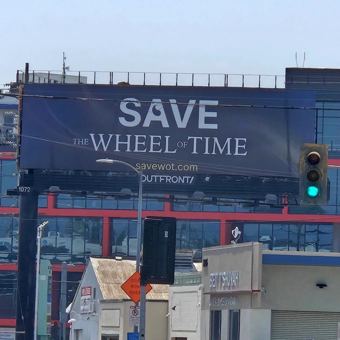 A photo of the big static billboard in Culver City with the text ¨Save the Wheel of Time¨ and the website savewot.com under it. In the letter A, part of the Save word is shown the sign of the female Aes Sedai organization - the flame of Tar Valon. The background is dark with visible waves of the One power like depicted in the Wheel of time TV show.