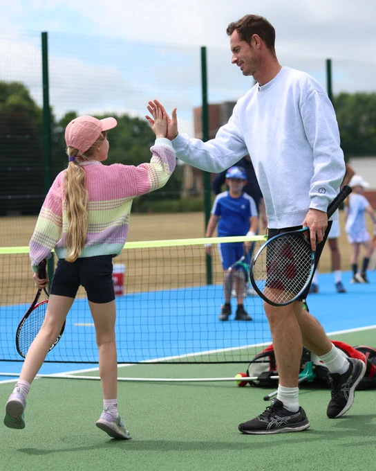 Photo of Andy Murray high-fiving a young girl on a park tennis court