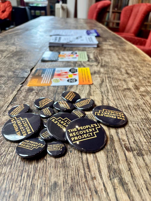 A photograph showing promotional badges for the 'The People's Recovery Project' and various leaflets all laid out on a table.
