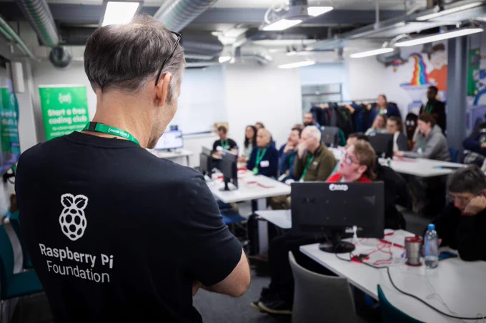 A photo of a person wearing a Raspberry Pi Foundation t-shirt standing at the front of a room full of people sitting at desks watching a talk at the front of a room. 