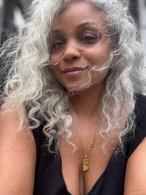 The author wearing a black top and gold necklace. Her white hair is blowing across her face.