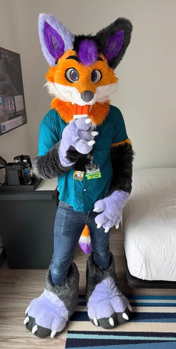 Photo of Scramble, a purple and orange fox fursuiter with mismatched ears, holding a fursuit fan that looks like a creamsicle 