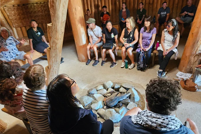 People sitting in a circle in an Indigenous building with cedar posts and walls.