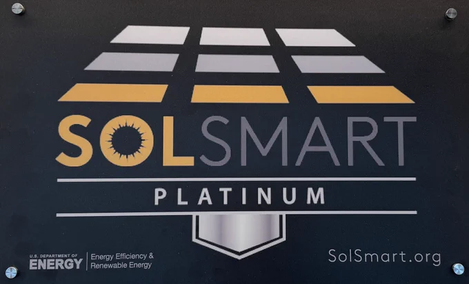 Plaque displaying the SolSmart Platinum designation. At the top is a stylized solar panel graphic in shades of gray and gold. Below, large text reads “SolSmart” with a sunburst design in the “O.” Underneath, a silver bar contains the word “Platinum.” In the lower left, it reads “U.S. Department of Energy, Energy Efficiency & Renewable Energy.” In the lower right: “SolSmart.org.”