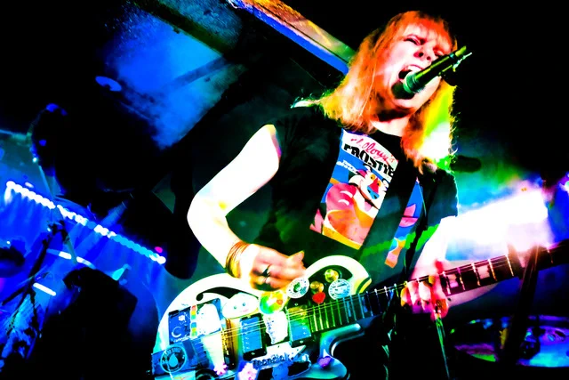 A person on stage, playing guitar and shouting into a microphone
