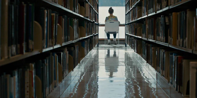 Looking up between two book stacks in a library towards a window where we can see the back of a woman sitting on a white plastic chair, working at a bench.