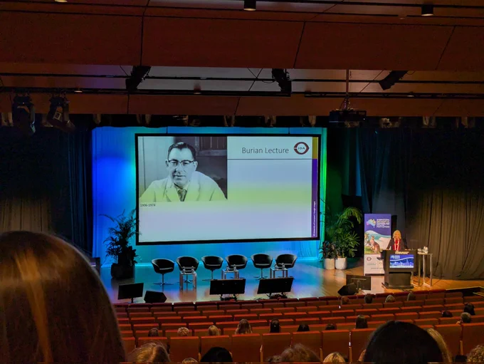 Fiona in a red top and black cardigan stands at the podium with a large screen on a stage in an auditorium