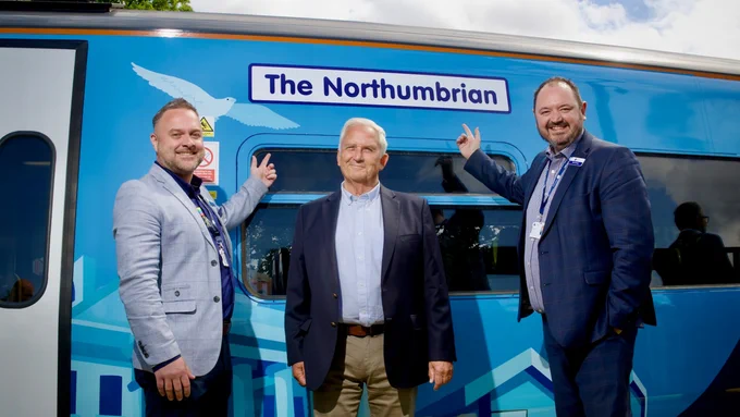 Three men standing in front of a blue train labeled "The Northumbrian." The men wear suits, with the two on the sides pointing at the train's name plaque. The train is decorated with a bird and a stylised landscape. The sky is partly cloudy.