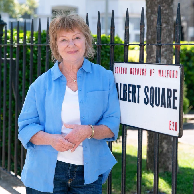 A woman stands by the railings in Albert Square, smiling. A street sign attached to the railings reads: London Borough of Walford, Albert Square, E20