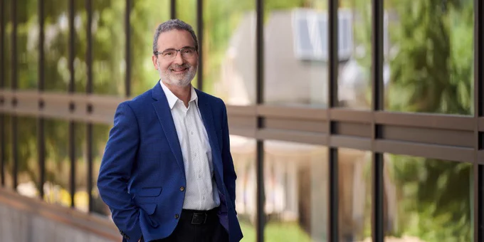 U of G president and vice-chancellor, Dr. Rene Van Acker stands in a blue blazer and white shirt smiling and standing in front of glass panels with trees in the background.
