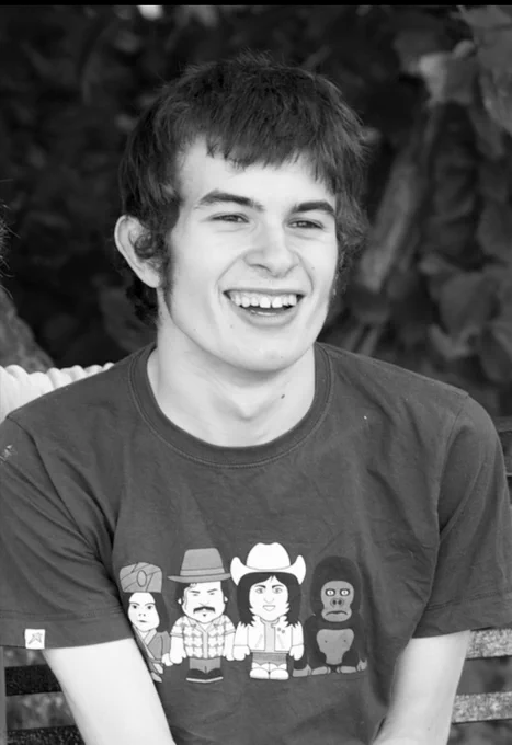 Black and white photo of Connor on a park bench wearing a Mighty Boosh t shirt. Massive smile.