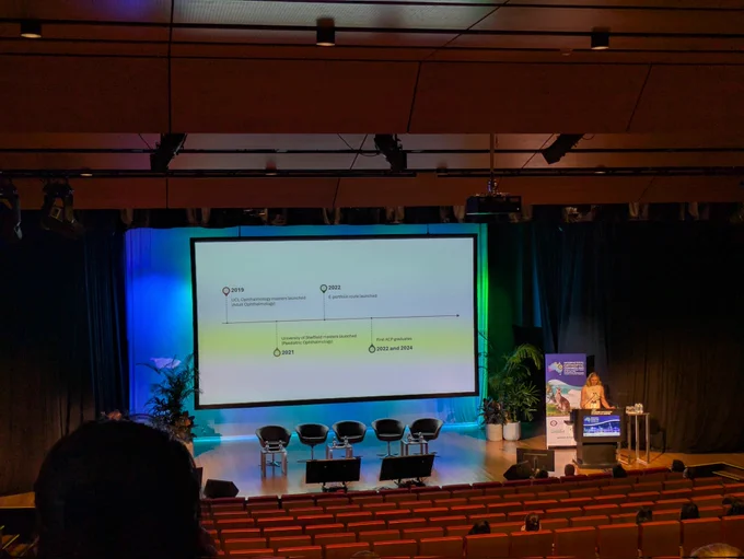 Kristina in a beige dress stands at the podium next to a large screen with slides