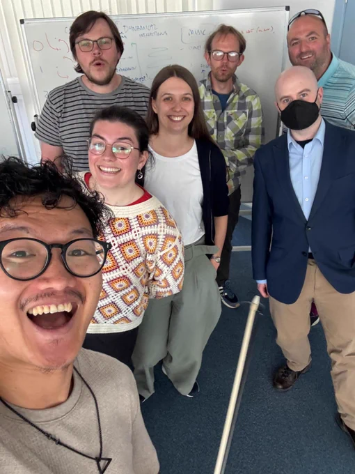 Photo of Science Ceilidh's Lewis developing the gravitational waves dance with scientists! Selfie of everyone together in front of a whiteboard, smiling. 7 people in photo 