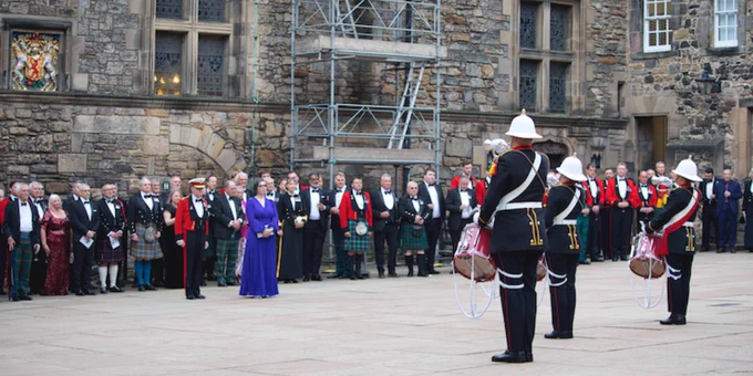 A group of people in formal attire, including military uniforms and traditional Scottish dress, watch a drumming performance by three Royal Marines drummers in ceremonial uniforms at Edinburgh Castle.
