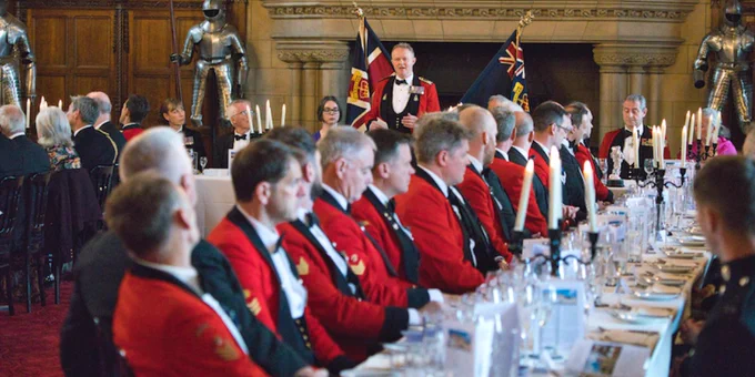Guests at a formal banquet at Edinburgh Castle, featuring individuals in traditional ceremonial red uniforms and others in formal wear, with a person standing and speaking at the head of the table, flanked by British flags.
