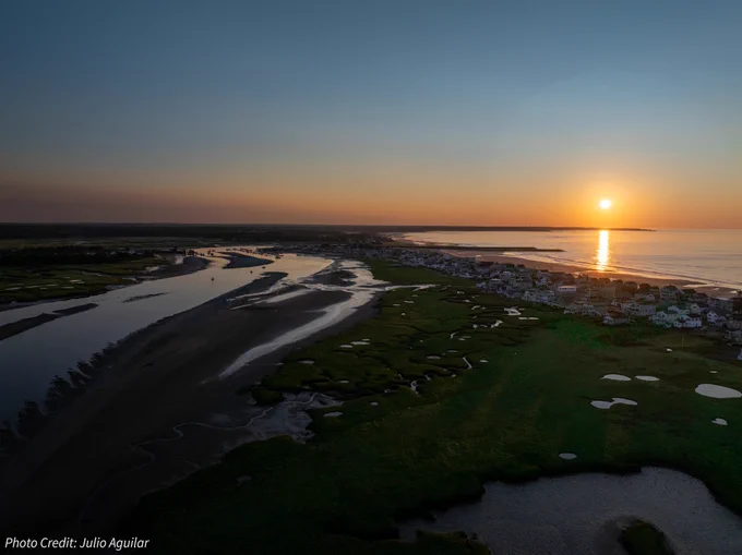 Aerial view of a marsh with a coastal town between it and the ocean at sunrise.