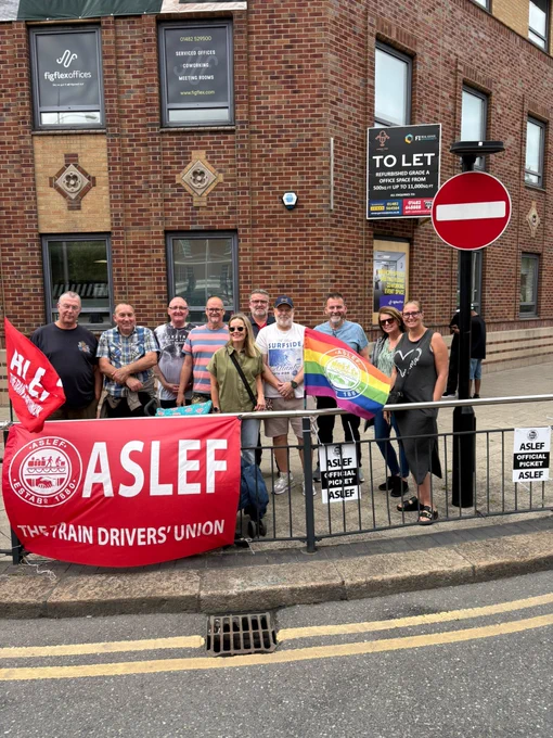 ASLEF Members standing on a picket line behind a road barrier with ASLEF flags and a Pride flag with the ASLEF logo on