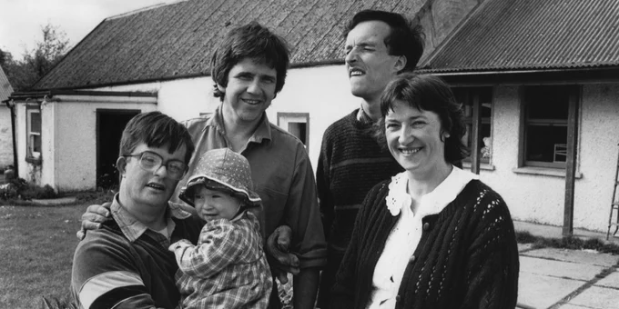 A black and white photo of Patrick and Gladys Lydon standing outside a white cottage with a corrugated roof.  Two other members of the community are standing beside them. One of them is holding a small child who is wearing a patterned bucket hat.