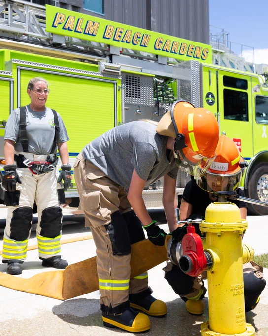 Two girls on fire camp attendees practice attaching a hose to a fire hydrant while a palm beach gardens firefighter oversees their work.