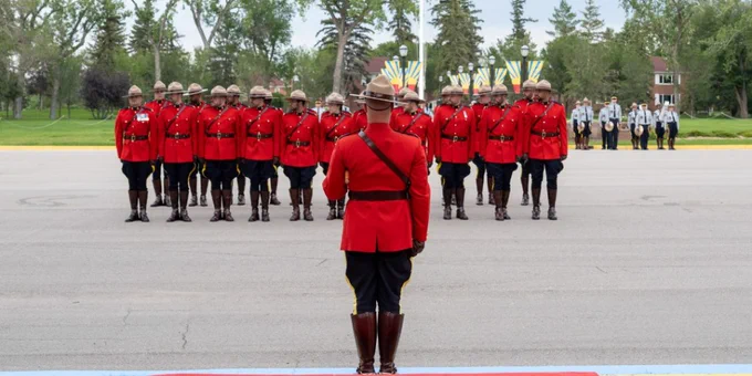 A troop of RCMP cadets in red serge stand together and look right. There is a drill instructor giving direction and there are members of the RCMP Cadet band and the public in the background.