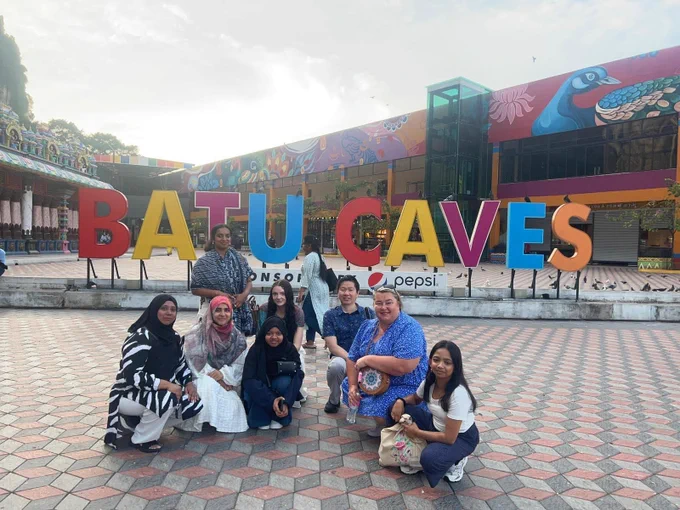 A group of eight people are posing in front of a large, colourful "BATU CAVES" sign. The letters of the sign are large, three-dimensional, and brightly coloured. Behind the sign, there is a building with murals, including a large peacock design. The ground is a patterned paved area. The sky is bright, suggesting daytime.