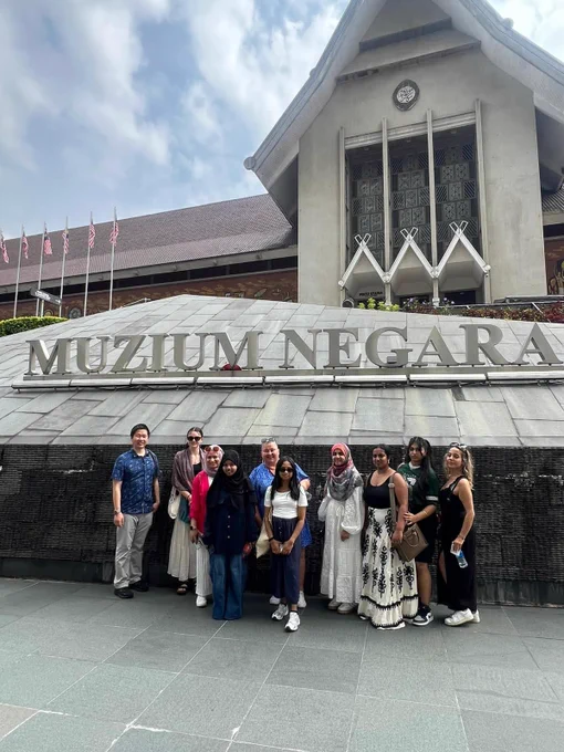 A group of ten people are standing in front of the "Muzium Negara" (National Museum) in Kuala Lumpur. The building behind them is a large, grey structure with traditional Malaysian architectural elements and a prominent sign displaying the museum's name. Several Malaysian flags are visible on flagpoles to the left. The group is smiling and posing for the camera, dressed in a variety of casual and traditional attire.