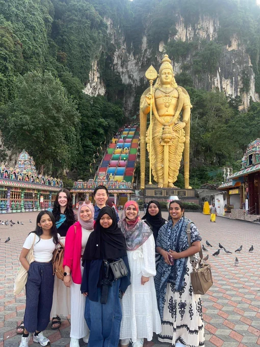 A group of eight individuals stands in the foreground, posing for a photo at the Batu Caves in Malaysia. Behind them, a massive golden statue of Lord Murugan towers over the scene. To the left of the statue, a vibrant, multi-coloured staircase ascends a steep natural rock formation. The ground is paved with pigeons scattered around. Traditional Hindu temple structures are visible on both sides of the foreground.