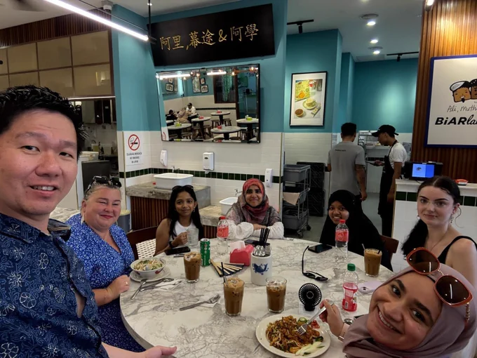 A group of eight people are seated around a circular white table in what appears to be a brightly lit restaurant or cafe. They are looking at the camera and smiling, with various drinks and plates of food on the table. The interior features light blue walls, white tiled lower walls, and mirrors reflecting parts of the room. Two staff members are visible in the background.