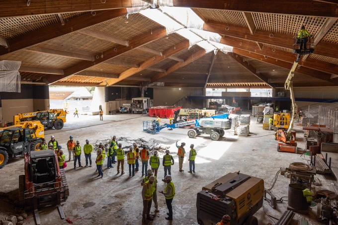 Interior view of a large construction site with Vail Town Council and others in safety vests gathered around. Construction equipment and materials are visible throughout the area. The ceiling features exposed wooden beams.